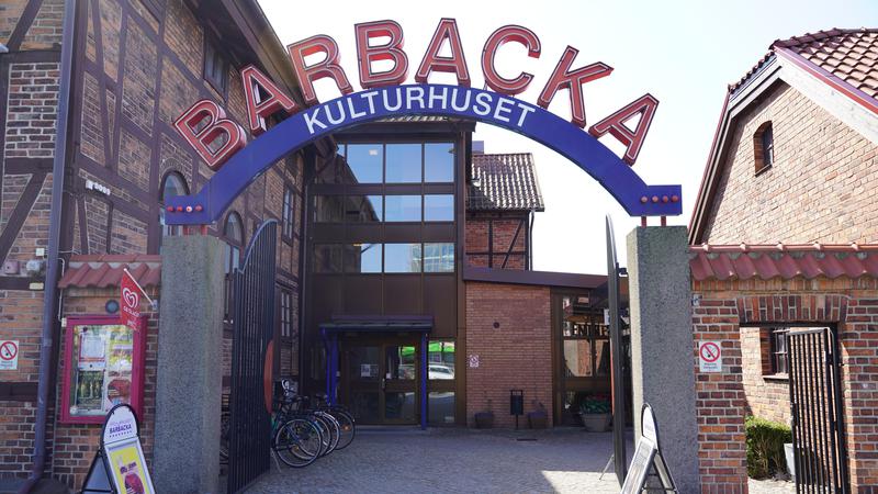 Två betongpelare håller uppe en välvd skylt i neon med orden "Barbacka Kulturhuset". I bakgrunden skymtar man entrén till byggnaden. 