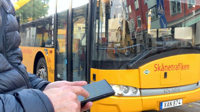 Resenär som kollar busstider i Skånetrafikens app, med gul regionbuss i bakgrunden